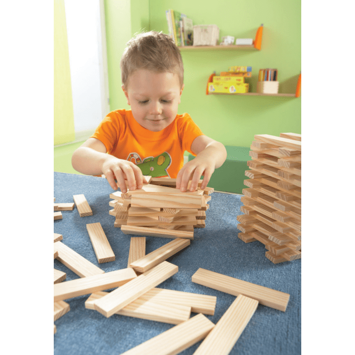 Ein kleines Kind in einem orangefarbenen Hemd baut ein Bauwerk aus Holzklötzen auf einem blauen Tisch, daneben ein weiterer Stapel Klötze und im Hintergrund Regale mit Büchern und Spielzeug.