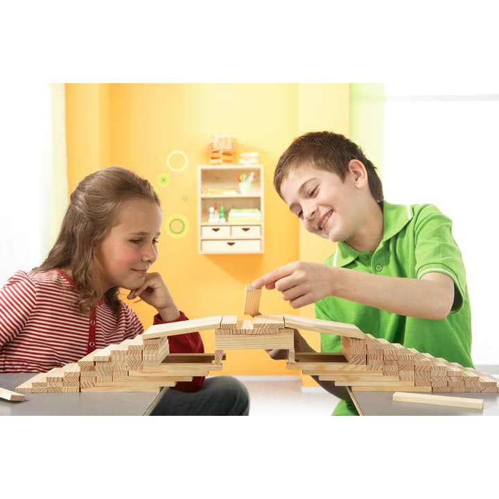 Ein Mädchen und ein Junge sitzen an einem Tisch und bauen gemeinsam eine Brücke aus Holzklötzen. Der Junge setzt den letzten Klotz oben auf, während das Mädchen zusieht. Der Hintergrund zeigt leuchtend grüne und orangefarbene Wände mit Regalen.