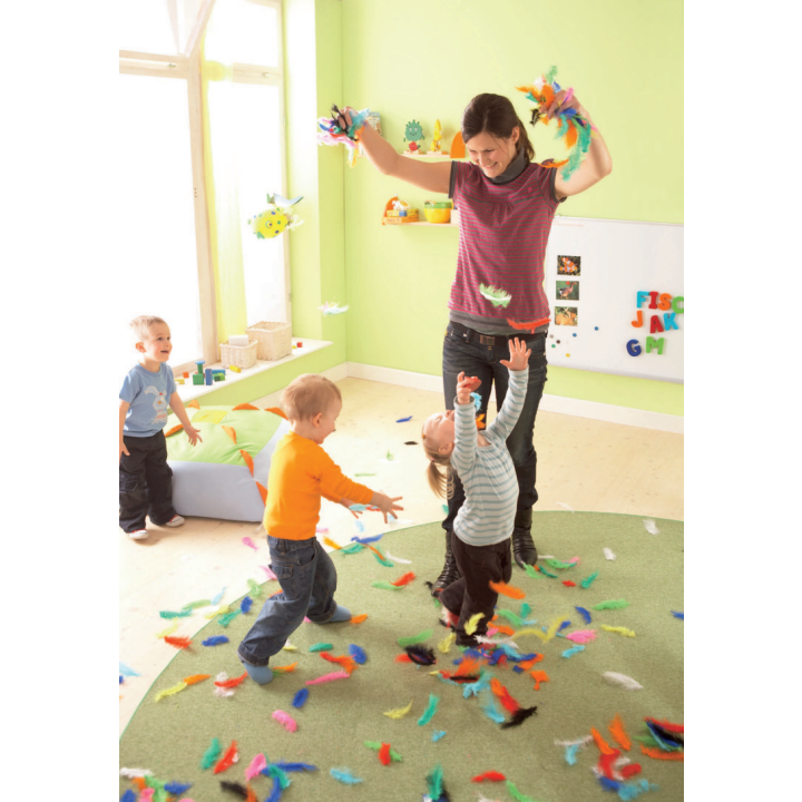 Ein Erwachsener und drei kleine Kinder spielen mit bunten Federn in einem hellen Raum. Die Federn sind auf dem Boden verstreut, und die Kinder greifen nach oben, während der Erwachsene die Federn in die Luft wirft.