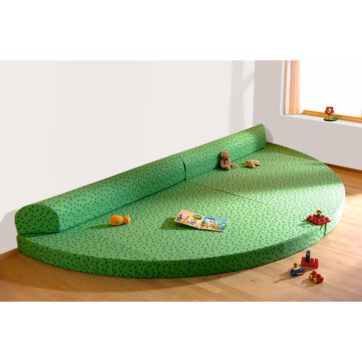 Eine große, grüne, halbrunde, gepolsterte Spielmatte mit einer Rückenlehne liegt in der Ecke eines Zimmers mit Holzboden. Kinderspielzeug und ein Buch liegen verstreut auf der Matte in der Nähe eines Fensters mit hellen Vorhängen.