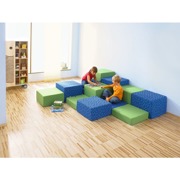 Zwei kleine Kinder sitzen auf grünen und blauen Schaumstoffblöcken, die als Sitzgelegenheiten in einem hellen Raum mit hellem Holzfußboden und blauen Wänden angeordnet sind. Im Hintergrund sind Regale mit Büchern und anderen Gegenständen zu sehen.