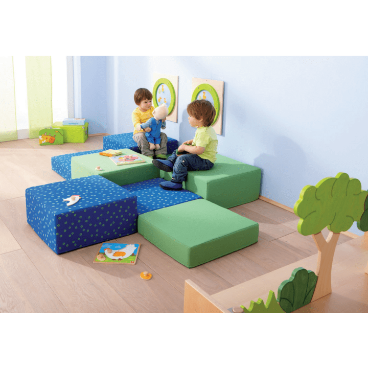 Zwei kleine Kinder sitzen auf grünen und blauen Schaumstoffblöcken in einem Spielzimmer und schauen sich eine Puppe an. Kinderbücher und Spielzeug liegen in der Nähe verstreut. Der Raum hat einen hellen Holzboden, blaue Wände und einfache Dekorationen.