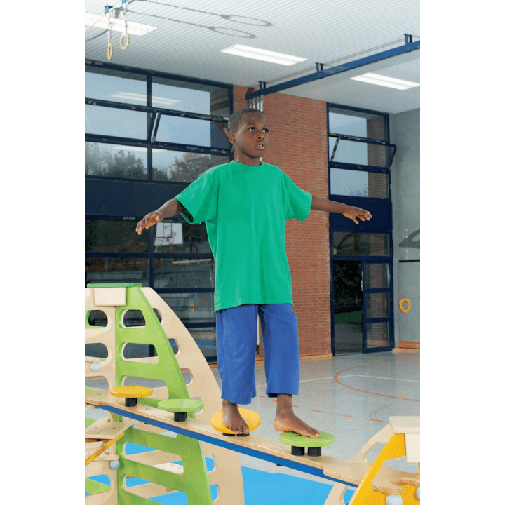 Ein Kind, das ein grünes T-Shirt und eine blaue Hose trägt, balanciert barfuß auf einem Holzbalken in einer Turnhalle, wobei es die Arme zum Gleichgewicht ausstreckt.