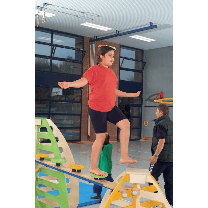 Ein Kind, das ein rotes Hemd, schwarze Shorts und ein grünes Stirnband trägt, balanciert auf einem Holzbalken in einer Turnhalle, während im Hintergrund Erwachsene und Kinder zu sehen sind.