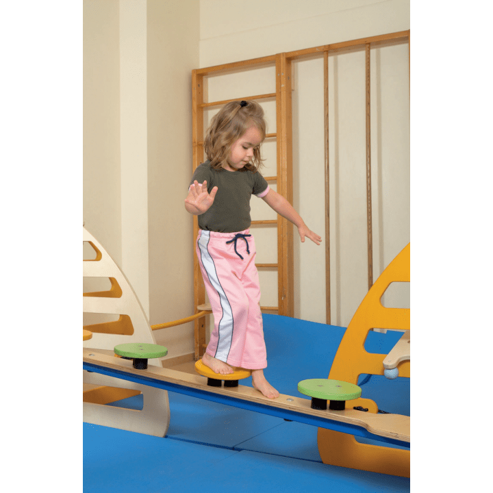 Ein kleines Kind mit hellbraunem Haar, das ein grünes T-Shirt und eine rosafarbene Hose trägt, balanciert barfuß auf einem Holzbrett in einem Indoor-Spielbereich mit blauen Matten und gelben Spielgeräten.