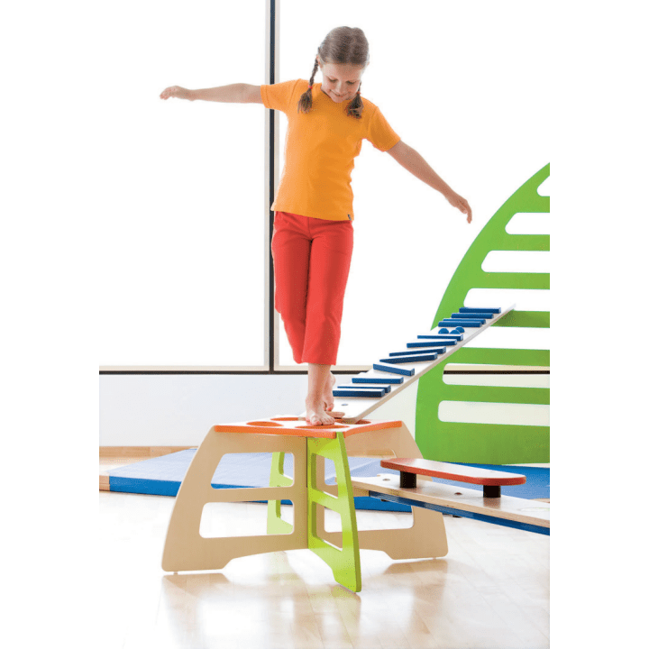 Ein junges Mädchen mit Zöpfen, das ein orangefarbenes Hemd und eine rote Hose trägt, balanciert barfuß auf einem hölzernen Spielgerät in einem Haus. Sie streckt ihre Arme aus, um das Gleichgewicht zu halten. Im Hintergrund sind eine blaue Matte und grüne Spielgeräte zu sehen.