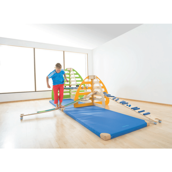 Ein Kind in blauem Hemd und roter Hose läuft auf einem schmalen hölzernen Balancierbalken, der an einem bunten Indoor-Spielgerät mit Klettergerüsten und blauen Matten auf einem hellen Holzboden befestigt ist.