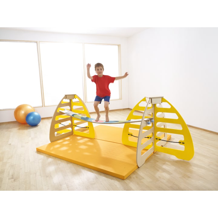 Ein kleines Kind in einem roten Hemd und blauen Shorts balanciert auf einer gebogenen Brettbrücke zwischen zwei gelben Klettergerüsten in einem hellen, geräumigen Raum mit großen Fenstern und Gymnastikbällen im Hintergrund.