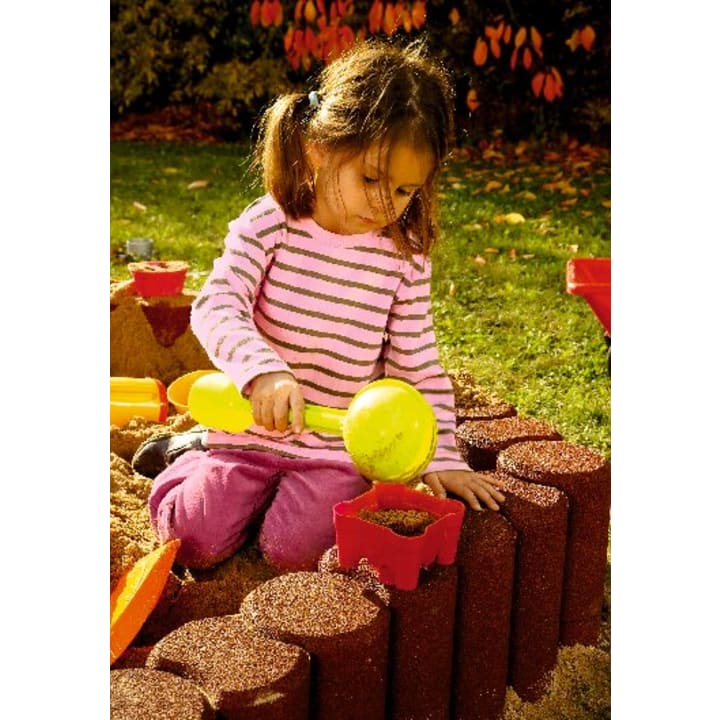 Ein junges Mädchen in einem rosa gestreiften Hemd und einer rosa Hose kniet in einem Sandkasten und benutzt eine gelbe Schaufel, um einen roten Behälter mit Sand zu füllen. Im Hintergrund sind Spielzeugblöcke und Herbstblätter zu sehen.