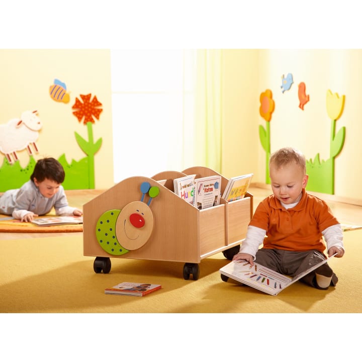 Zwei kleine Kinder befinden sich in einem bunten Spielzimmer. Ein Kind sitzt und schaut sich ein Buch neben einem Bücherwagen in Form einer Raupe an. Ein anderes Kind liegt mit einem Buch auf dem Boden im Hintergrund. Helle Wanddekorationen sind zu sehen.