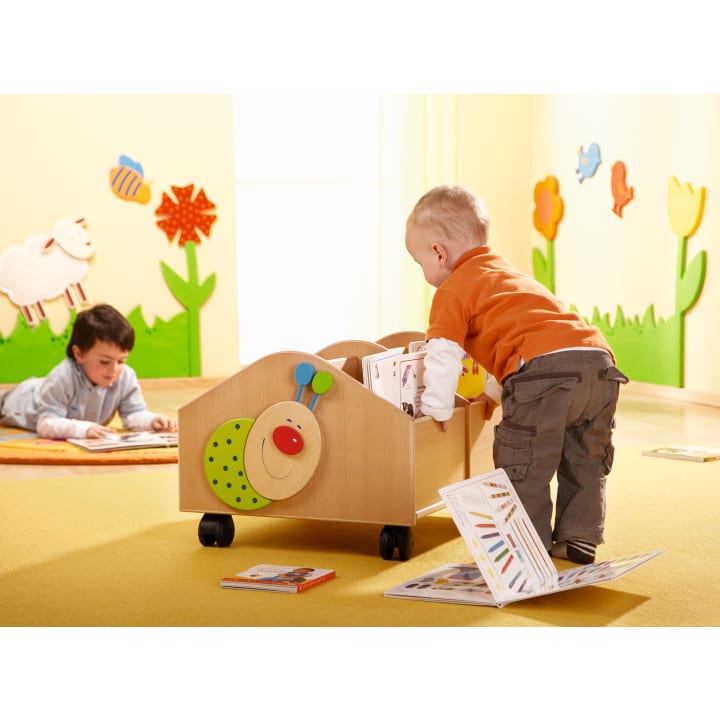 Zwei kleine Kinder befinden sich in einem bunten Spielzimmer. Ein Kind steht in einem hölzernen Aufbewahrungswagen in Form einer Raupe und blättert in Büchern, während das andere Kind auf dem Boden liegt und ein Buch liest.