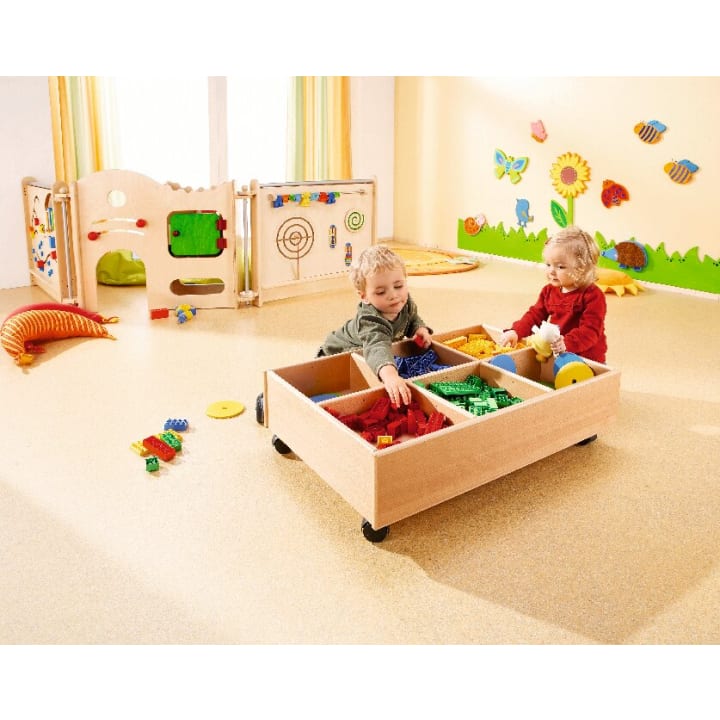 Zwei kleine Kinder sitzen auf dem Boden eines hellen, organisierten Spielzimmers und spielen mit bunten Bauklötzen aus einem hölzernen Aufbewahrungswagen. Das Zimmer hat Wanddekorationen und verschiedene Spielzeuge im Hintergrund.