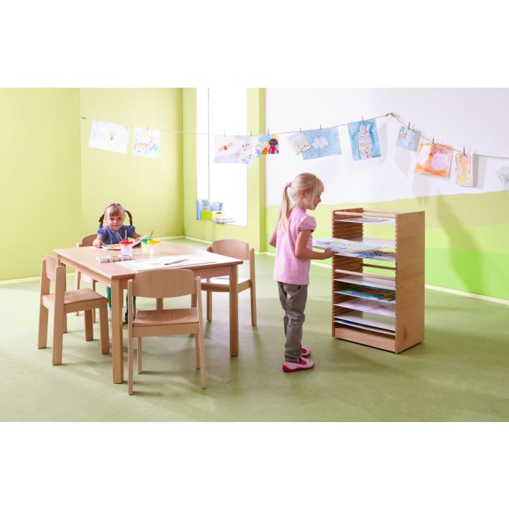 Zwei junge Mädchen befinden sich in einem hellen Klassenzimmer mit grünem Boden und grünen Wänden. Ein Mädchen sitzt an einem Tisch mit Malutensilien, während das andere seine Kunstwerke auf einen hölzernen Trockenständer legt. Fertige Zeichnungen hängen an einer Schnur quer durch den Raum.