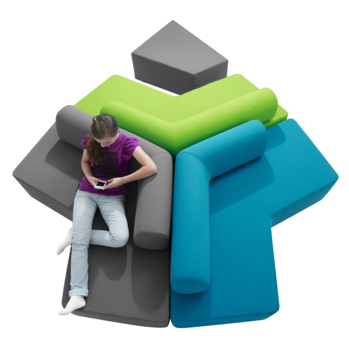 Eine Person sitzt auf einem modularen, sternförmigen Sofa mit grauen, grünen und blauen Teilen und zylindrischen Rückenlehnen und schaut auf ein Smartphone. Das Sofa steht vor einem weißen Hintergrund.