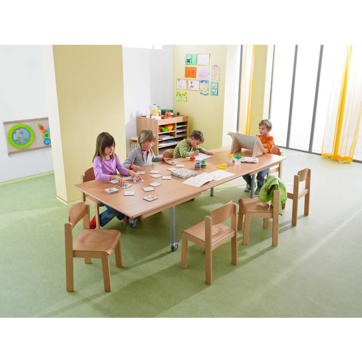 Vier kleine Kinder sitzen an einem Holztisch in einem Klassenzimmer und beschäftigen sich mit verschiedenen künstlerischen und handwerklichen Aktivitäten. Auf dem Tisch liegen Malutensilien, und an den Wänden hängen die Kunstwerke der Kinder. Der Raum ist von hellem Tageslicht durchflutet.