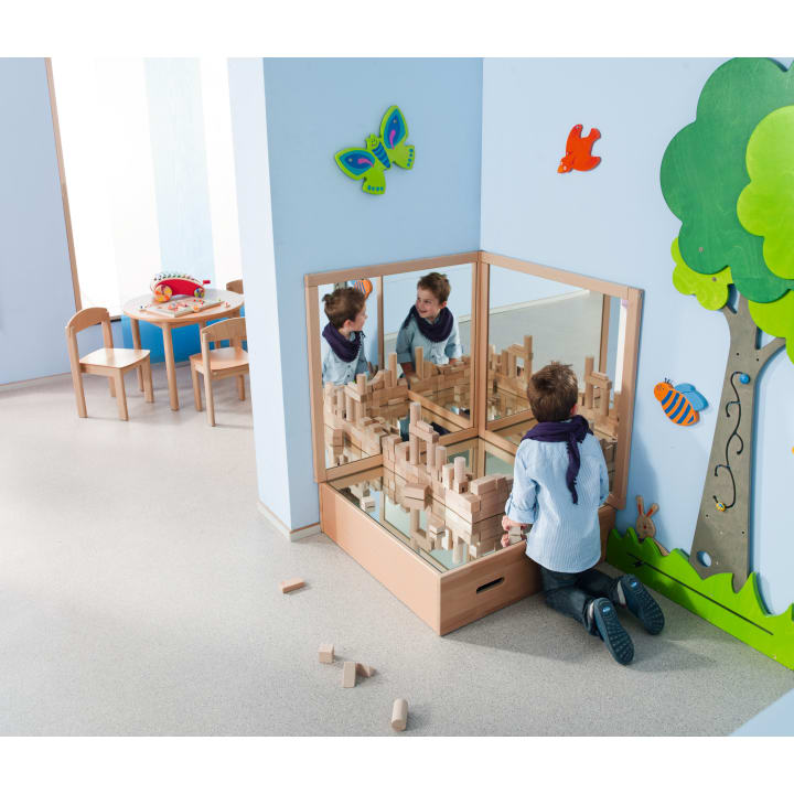 Ein Kind kniet vor einer mit Holzklötzen gefüllten Spiegelecke und baut ein Bauwerk. Im Hintergrund sind bunte Dekorationen, ein Tisch und Stühle zu sehen.