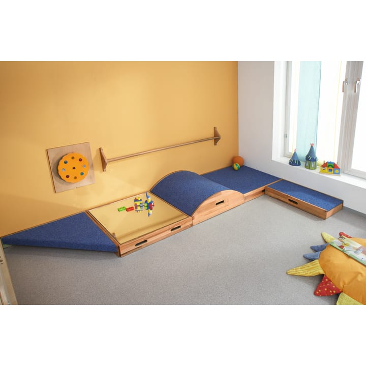 Ein Kinderspielzimmer mit einer gelben Wand, blauen und hölzernen Spielstrukturen, einem an der Wand befestigten runden Puzzle, verstreutem Spielzeug, einer gepolsterten Matte und weichen Kissen in Form von Sonnenstrahlen auf einem hellgrauen Boden.