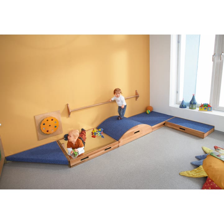 Zwei kleine Kinder spielen auf niedrigen, weichen Klettergerüsten in einem hell erleuchteten Raum mit gelben Wänden, einem Fenster und buntem Spielzeug, das überall verstreut ist. Ein Kind läuft auf einer erhöhten Plattform, während das andere in der Nähe krabbelt.