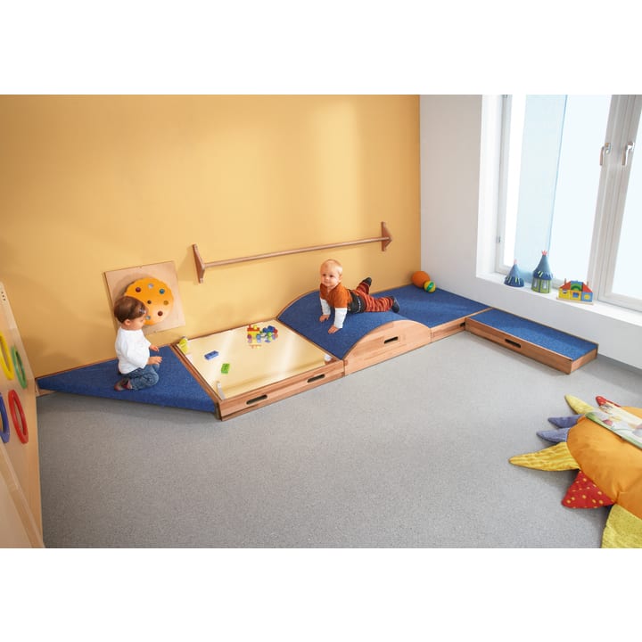 Zwei Kleinkinder spielen in einem Raum mit einer gelben Wand und einem blauen, gepolsterten Spielgerät. Ein Kind sitzt auf der linken Seite und spielt mit Spielzeug, während das andere auf der rechten Seite krabbelt. Weiche Matten und bunte Spielsachen liegen verstreut herum.