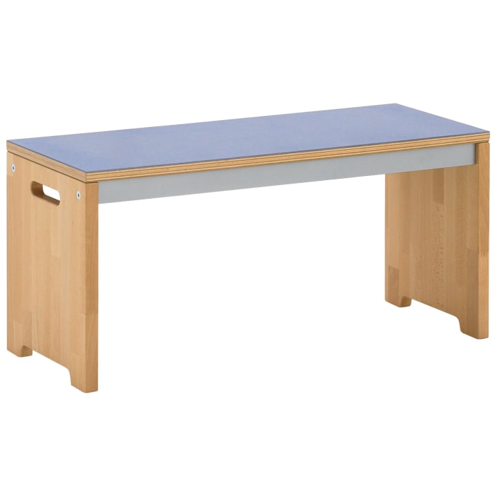 Eine Holzbank mit hellen Beinen und einer flachen, blauen Sitzfläche. Die Bank hat einfache, rechteckig ausgeschnittene Griffe auf jeder Seite und ein minimalistisches, modernes Design.