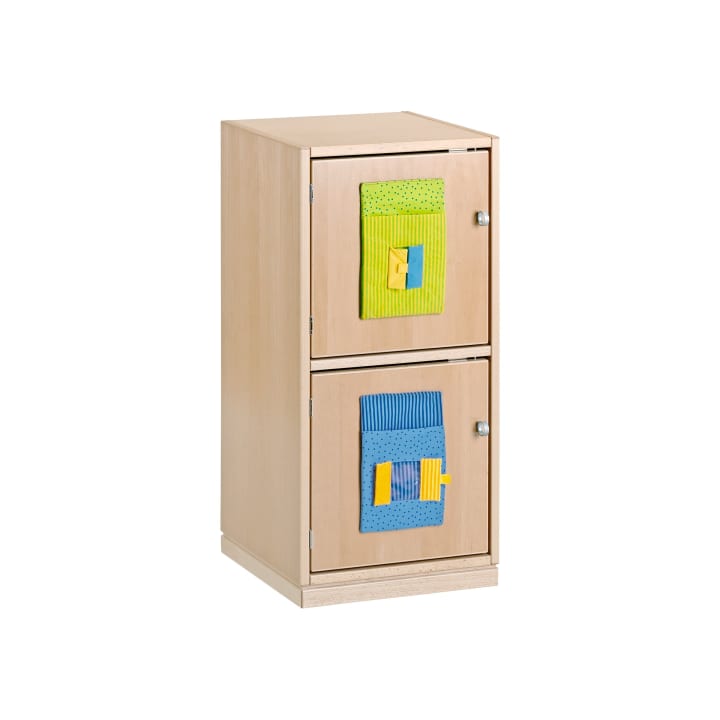 Ein Aufbewahrungsschrank aus hellem Holz mit zwei Fächern, die jeweils mit einem Stoffquadrat an der Tür versehen sind. Das obere Quadrat ist grün und gelb, das untere ist blau und gelb. Beide haben ein strukturiertes Muster und kleine Griffe.
