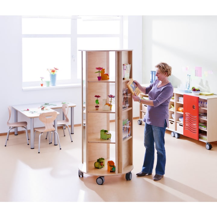 Eine Person steht neben einem hohen, drehbaren Holzregal auf Rädern in einem hellen Klassenzimmer und ordnet Bücher ein. Der Raum hat Tische und Stühle in Kindergröße und Schränke mit Pflanzen und Dekorationen.