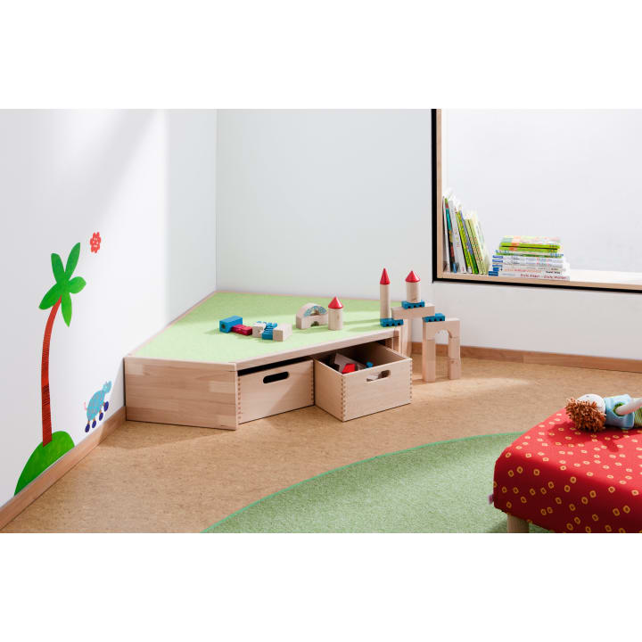 Eine Kinderspielecke mit einem Holzregal mit Bauklötzen, einer roten Bank mit einer Puppe, einem grünen Teppich und einer Wand mit einer Palme und Büchern am Fenster.