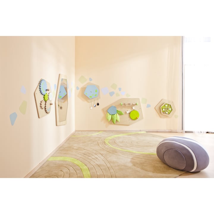 Ein Spielzimmer mit hellbeigen Wänden, interaktiven Holzpaneelen mit grünen und blauen Elementen an einer Wand, einem beigen Teppich mit grünen Wirbeln und einem weichen, abgerundeten grauen und weißen Sitz. Natürliches Licht kommt von einem großen Fenster.