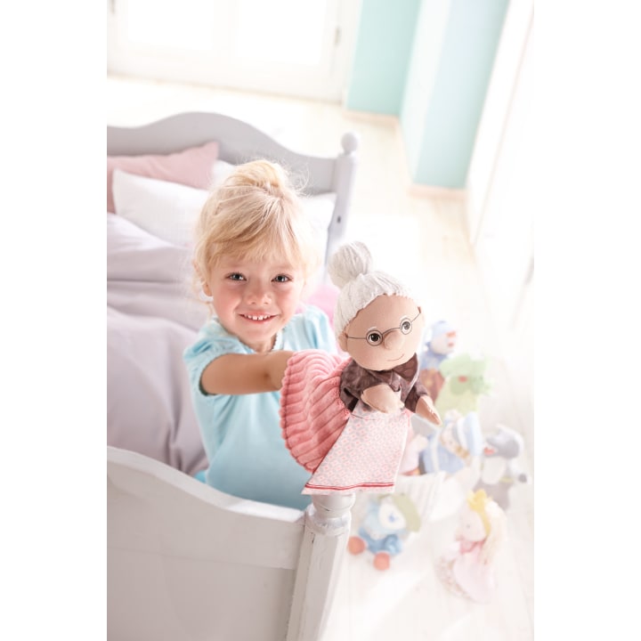 Ein kleines Kind mit blondem Haar steht lächelnd an einem weißen Bett und hält eine Puppe hoch, die wie eine ältere Frau mit Brille und grauem Haar aussieht. Im Hintergrund sind mehrere Plüschtiere und Spielzeuge zu sehen.