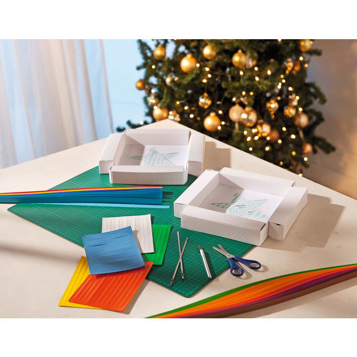 Ein Tisch mit bunten Quilling-Papierstreifen, zwei laufenden Quilling-Projekten auf weißen Tabletts, Pinzetten, Scheren und Bastelwerkzeug. Im Hintergrund steht ein geschmückter Weihnachtsbaum mit Ornamenten.
