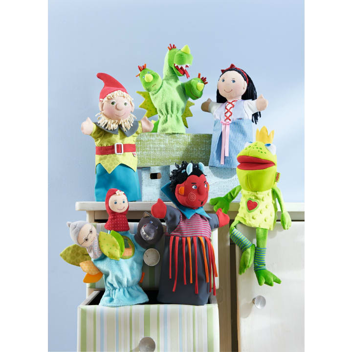 Sechs bunte Handpuppen, darunter ein Gnom, ein grüner Drache, ein Mädchen in einem blauen Kleid, ein Frosch, eine Hexe und eine Figur mit einem roten Umhang, sind auf Schubladen und Kisten vor einer hellblauen Wand angeordnet.