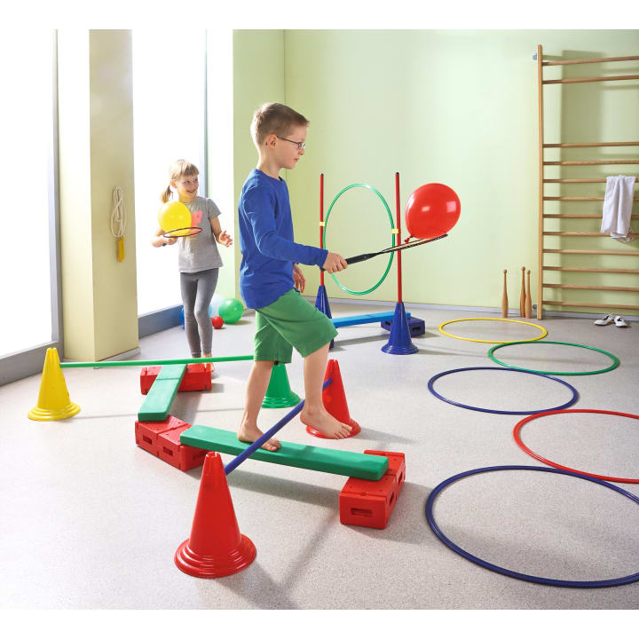 Zwei Kinder spielen in einem Turnraum mit bunten Kegeln, Reifen und Schwebebalken. Der Junge läuft barfuß auf einem Balken und balanciert einen roten Luftballon auf einem Stock, während das Mädchen mit einem gelben Ball hinterherläuft.