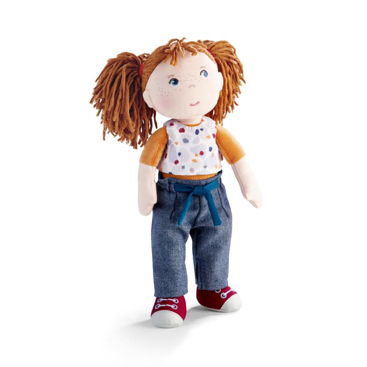 Eine Stoffpuppe mit rotem Garnhaar in Zöpfen, die ein weißes Hemd mit bunten Flecken und orangefarbenen Ärmeln, blaue Jeans mit blauem Gürtel und rote Turnschuhe trägt, steht vor einem weißen Hintergrund.