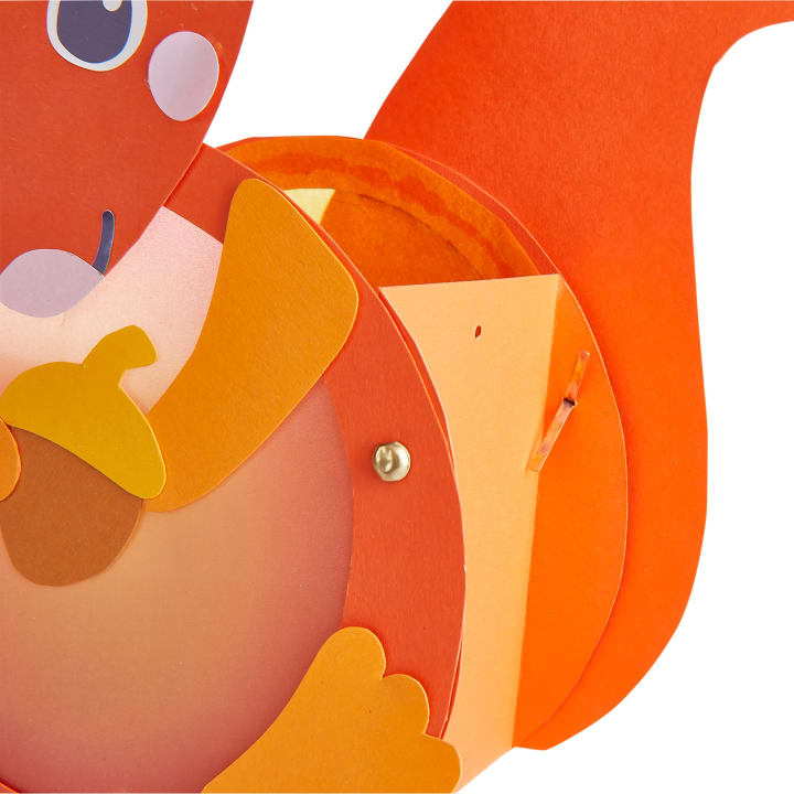 Nahaufnahme einer bunten orange-roten Papierarbeit in Form eines Eichhörnchens, das eine gelbe und braune Eichel hält. Die Bastelarbeit besteht aus mehreren Schichten von Papierstücken und einem sichtbaren Metallverschluss.