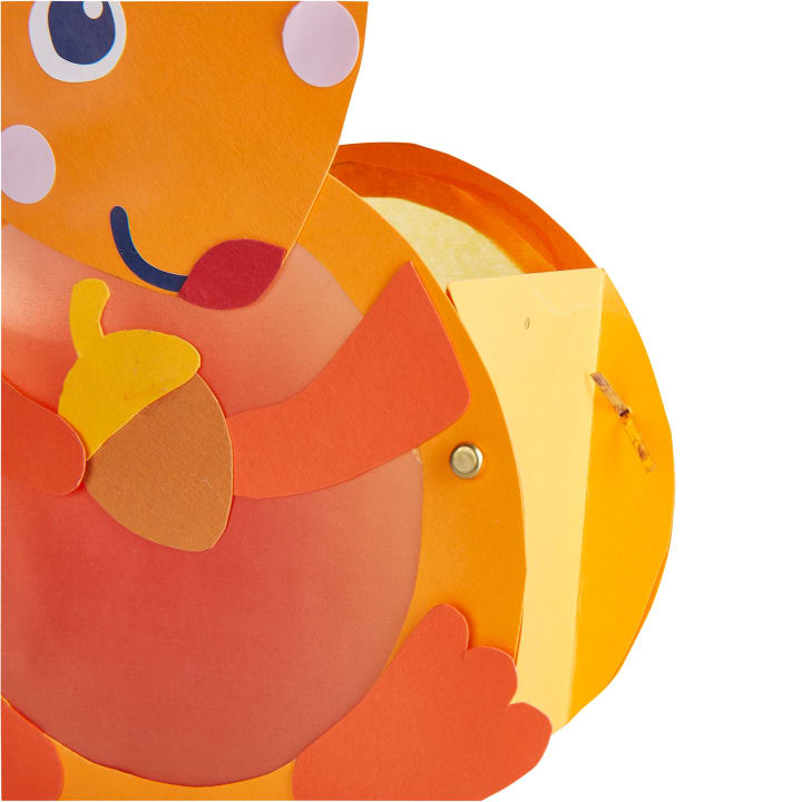 Eine Nahaufnahme einer bunten, orangefarbenen, eichhörnchenförmigen Papierlaterne, die eine gelbe Eichel hält, mit sichtbaren geschichteten Papierstücken und einem Metallverschluss an der Seite.