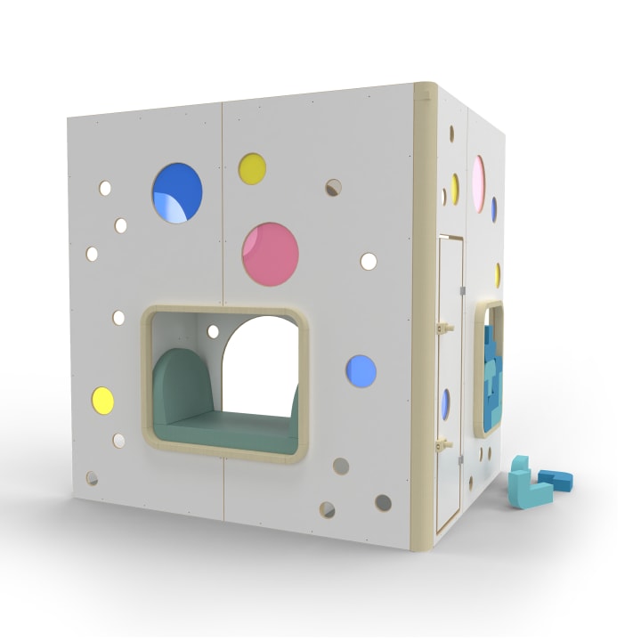 Ein weißes, kubisches Spielhaus mit kreisförmigen Ausschnitten in verschiedenen Farben, einer Fensterbank im Inneren und Schaumstoffblöcken auf dem Boden in der Nähe. Die Struktur hat ein modernes Design und scheint für Kinder gedacht zu sein.