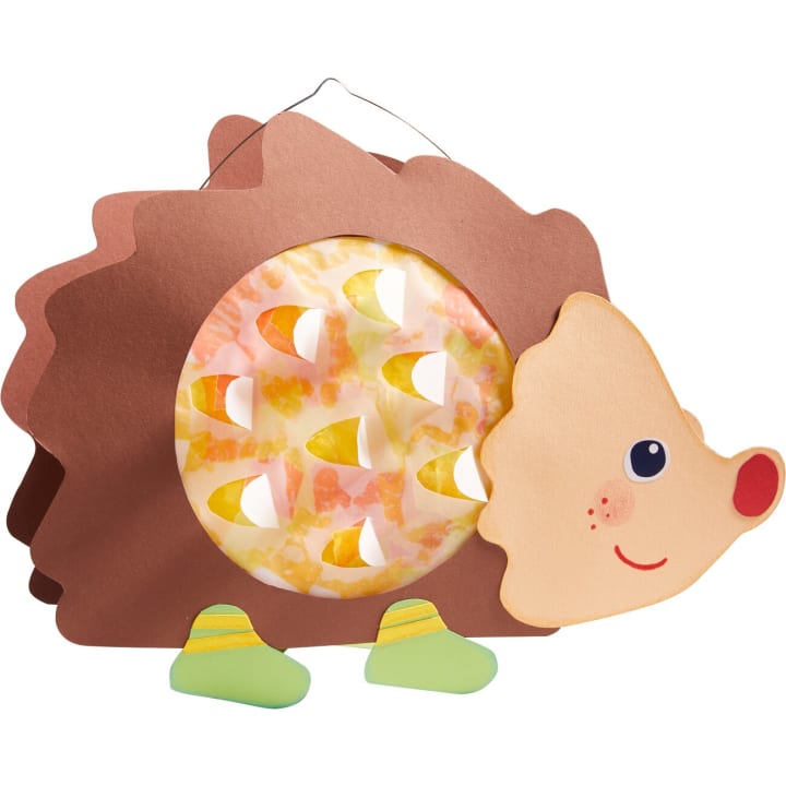 Ein handgefertigter Igel aus braunem und beigem Papier mit einer runden, durchsichtigen Mitte, die mit buntem Seidenpapier und kleinen ausgestanzten Formen gefüllt ist. Der Igel hat Papierfüße, eine rote Nase und ein gezeichnetes Lächeln.