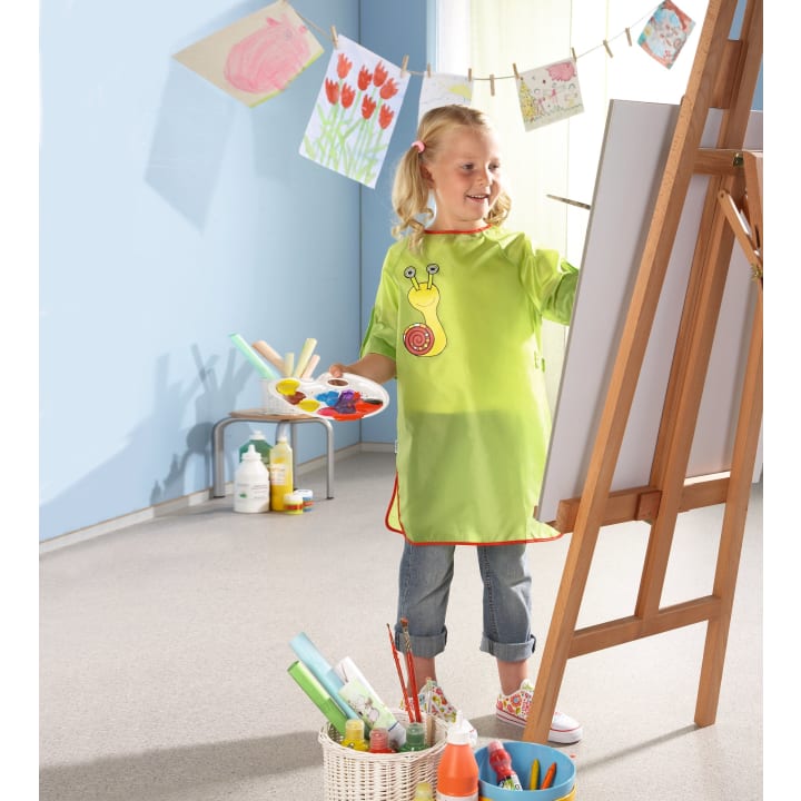Ein junges Mädchen in einem grünen Kittel steht an einer Staffelei und malt auf einer Leinwand. In einem Korb auf dem Boden liegen Malutensilien, und im Hintergrund hängen Kinderzeichnungen an einer Wäscheleine.