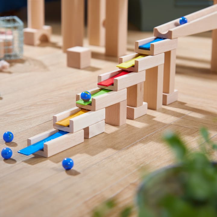 Auf einer Holzfläche ist eine hölzerne Murmelbahn mit Rampen in den Farben Blau, Gelb und Rot aufgebaut. Mehrere blaue Murmeln befinden sich auf und um die Struktur herum. Eine unscharfe grüne Pflanze erscheint in der unteren rechten Ecke.