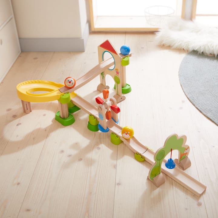 Auf einem hellen Holzboden ist eine Holzmurmelbahn mit Rampen, einer gelben Spiralbahn, bunten Stützen und dekorativen Elementen wie Bäumen und kleinen Figuren mit aufgemalten Gesichtern aufgebaut.