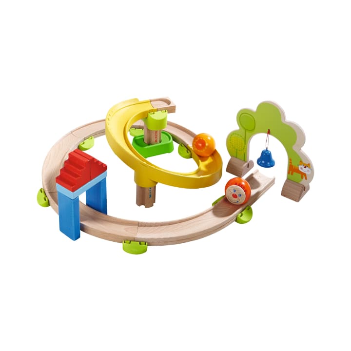 Holzmurmelbahn mit kreisförmiger Bahn, gelber Spiralrampe, roter und blauer Treppe, einer Glocke, grünen Verbindungsstücken und einer baumförmigen Platte. Eine orangefarbene Kugel und eine runde Figur mit Tiergesicht sind ebenfalls enthalten.