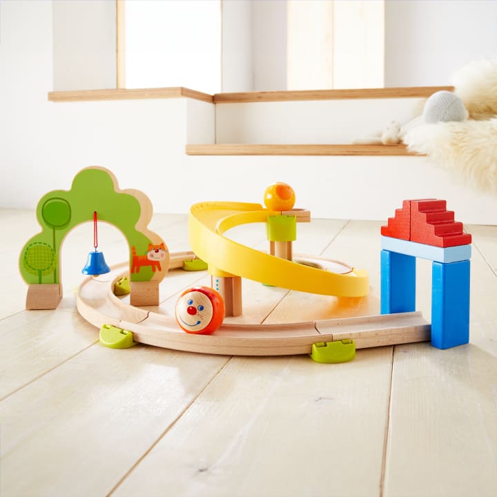 Ein Spielzeugeisenbahnset aus Holz mit einer spiralförmigen gelben Rampe, einer Kugel mit einem lächelnden Gesicht, einer blauen und roten Hausstruktur und grünen baumförmigen Bögen auf einem hellen Holzboden in einem hellen Raum.