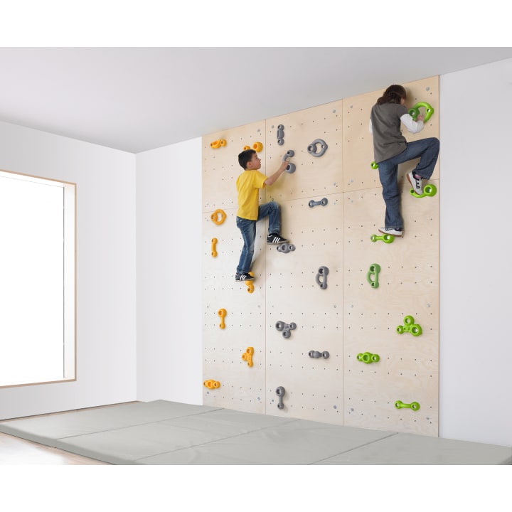 Zwei Kinder klettern an einer Indoor-Kletterwand aus Holz mit bunten Griffen. Die Wand befindet sich in einem hellen Raum mit einem großen Fenster und einer gepolsterten Matte auf dem Boden zur Sicherheit.