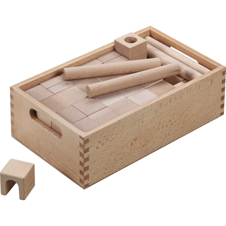 Eine Holzkiste enthält verschiedene Holzklötze, Zylinder und ein kleines Bogenstück aus hellem Holz. Einige Klötze sind ordentlich im Inneren der Kiste angeordnet, während andere oben oder außen platziert sind.
