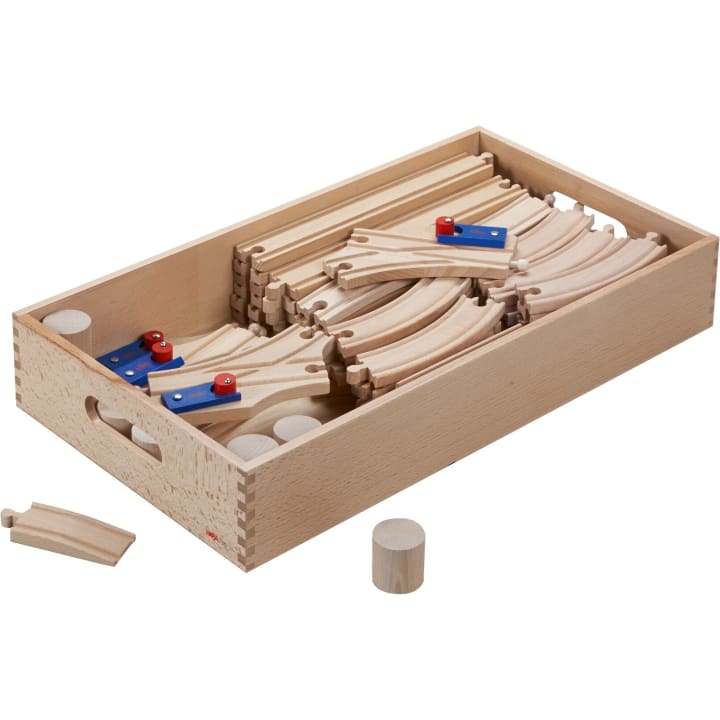 Eine Holzkiste mit verschiedenen Holzschienen und Verbindungsstücken, mit einigen gebogenen und geraden Abschnitten und zwei zylindrischen Teilen. Ein gebogenes Gleis und ein zylindrisches Stück befinden sich außerhalb der Kiste.