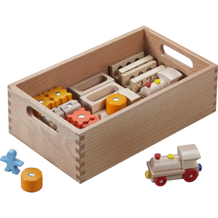 Eine Holzkiste mit verschiedenen Holzspielzeugteilen, darunter Zahnräder, Blöcke und zylindrische Teile. Eine kleine zusammengebaute Holzeisenbahn und eine blaue Figur befinden sich außerhalb der Kiste auf weißem Hintergrund.