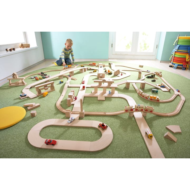 Ein kleines Kind sitzt auf einem grünen Teppich und spielt mit einer großen Holzeisenbahn und einem Straßenset mit verschiedenen kleinen Spielzeugautos und -fahrzeugen, die auf den Gleisen und Strukturen angeordnet sind. Im Hintergrund stehen gestapelte bunte Stühle.