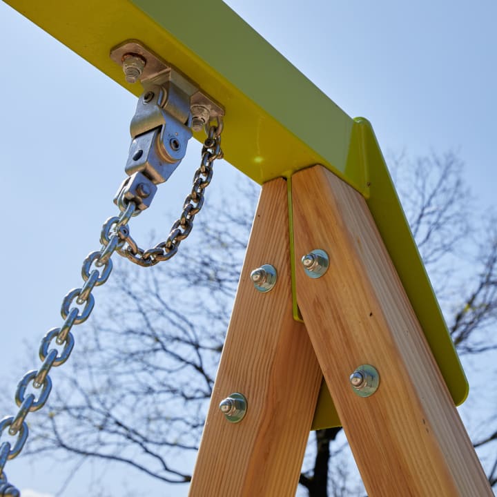 Nahaufnahme einer Metallkette und einer Halterung, die an einem hölzernen A-Rahmen befestigt sind, wahrscheinlich Teil einer Schaukel, mit Schrauben zur Sicherung des Rahmens und einem klaren Himmel im Hintergrund.