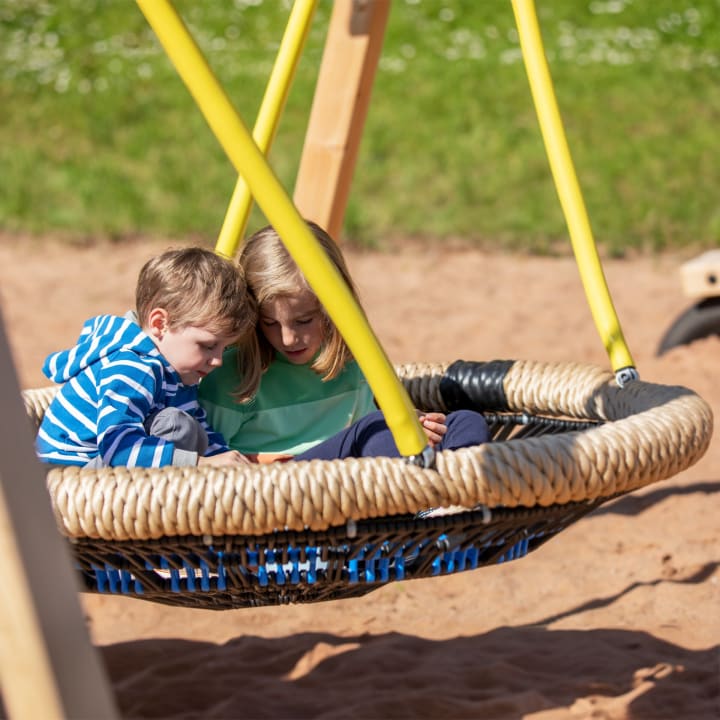 Zwei kleine Kinder sitzen zusammen auf einer großen runden Seilschaukel auf einem Spielplatz. Die Schaukel hängt an gelben Seilen über einer sandigen Fläche, und beide Kinder schauen nach unten, möglicherweise auf etwas in ihrem Schoß.