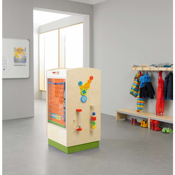 Eine hölzerne Spielstation steht in der Mitte eines Raums mit grauen Böden und Wänden. Auf der rechten Seite befindet sich eine Garderobe mit bunten Kinderjacken, Stiefeln und Schuhen. Im Hintergrund befinden sich ein Whiteboard und ein Fenster.